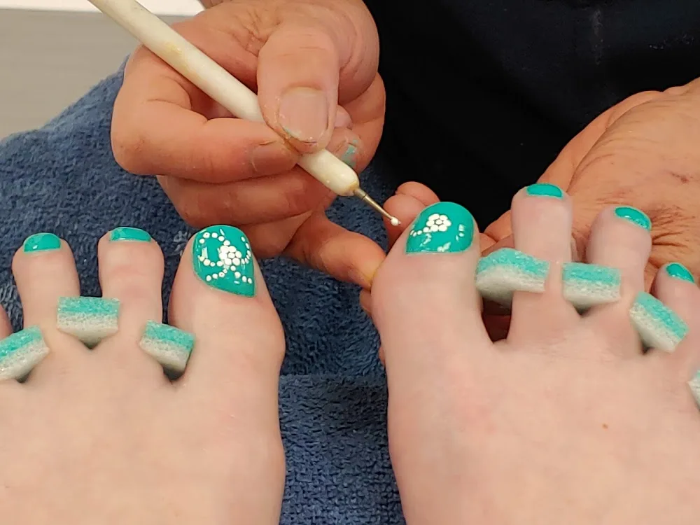 A technician hand painting white flowers onto a teal nail with a dotting tool